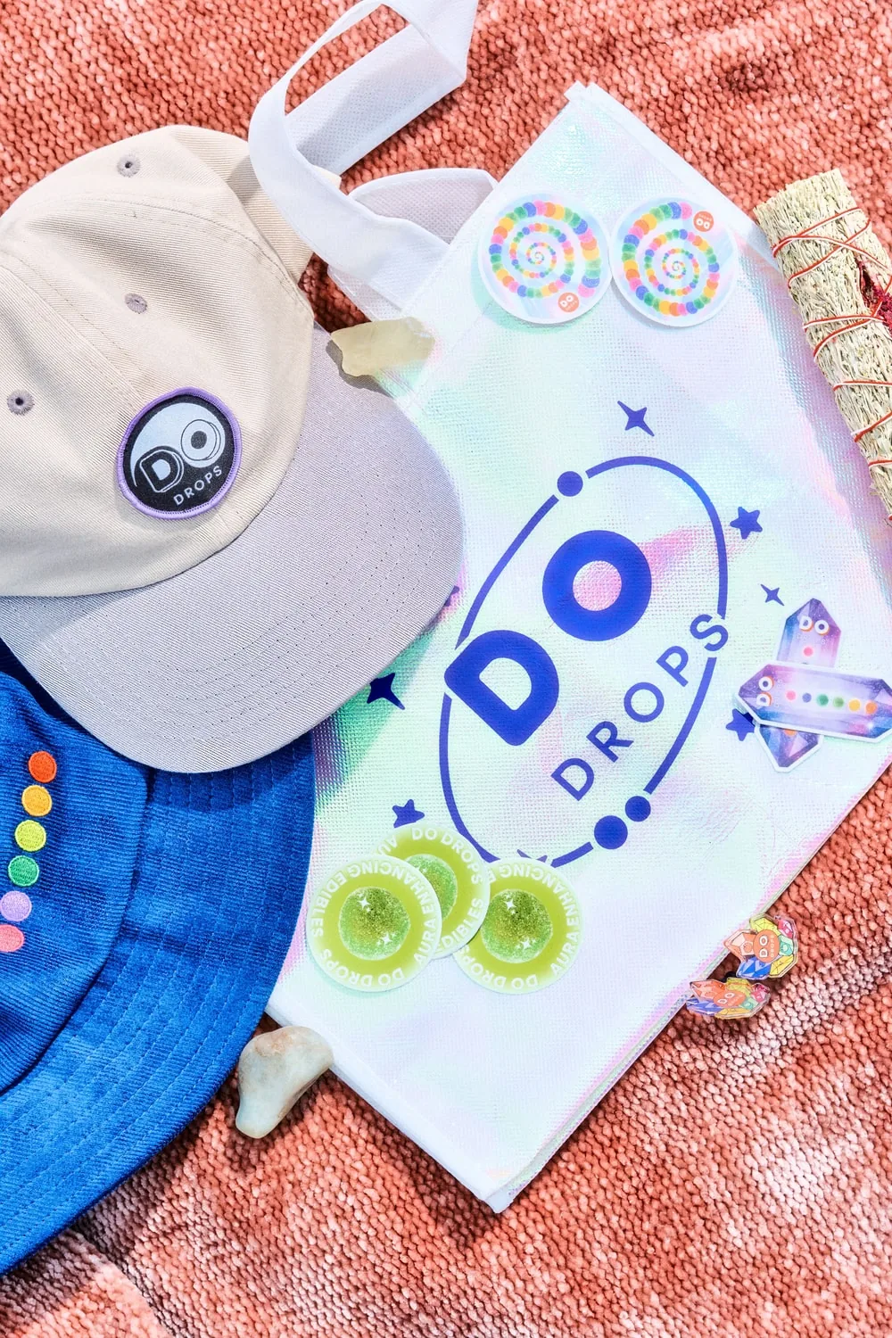 DO Drops merchandise flatlay: an embroidered cap, an iridescent tote, enamel pins and stickers arranged on a woven blanket.