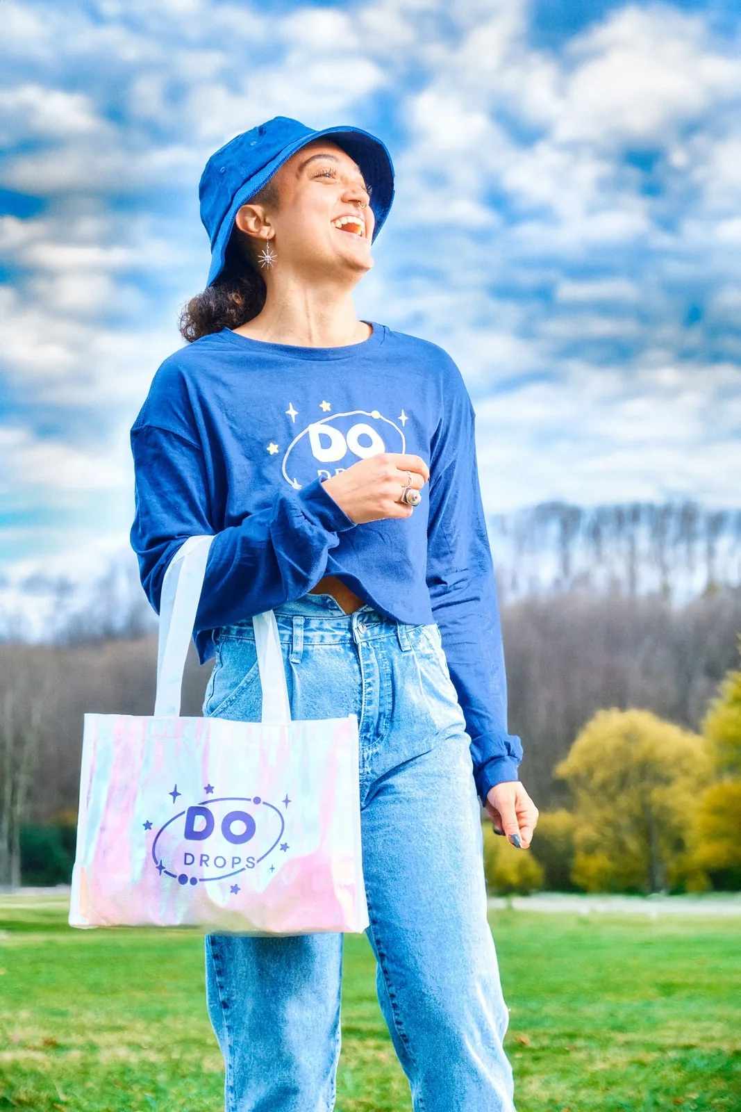 A model in a blue DO Drops long-sleeve and bucket hat carrying an iridescent DO Drops tote in an open field.