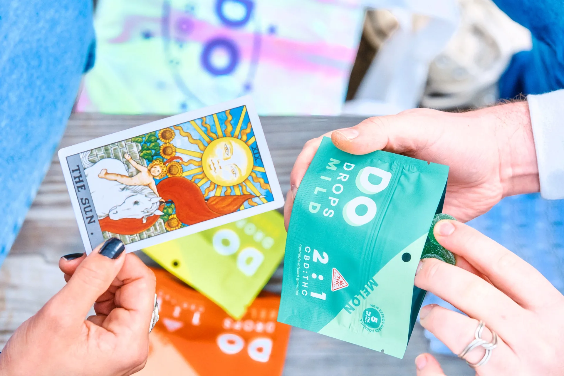 Hands holding a DO Drops Mild pouch beside a brightly illustrated tarot card on a wooden table.